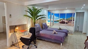 A bedroom with two beds, a desk, a chair, a large plant, and a view of the beach through the windows. - T2 45m2 with garden, Air conditioning, Parking, Quiet, Comfortable, Beach 800m (Saint-Raphaël)