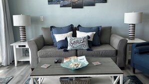 Ein Wohnzimmer mit grauem Sofa, zwei Beistelltischen mit Lampen, einem Couchtisch mit Fernbedienungen und einem Korb sowie einer Wand mit einem mehrteiligen Kunstwerk.