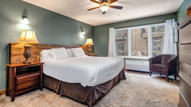 Une chambre avec un grand lit, deux lampes de chevet, un ventilateur de plafond, une fenêtre avec des rideaux et un fauteuil.