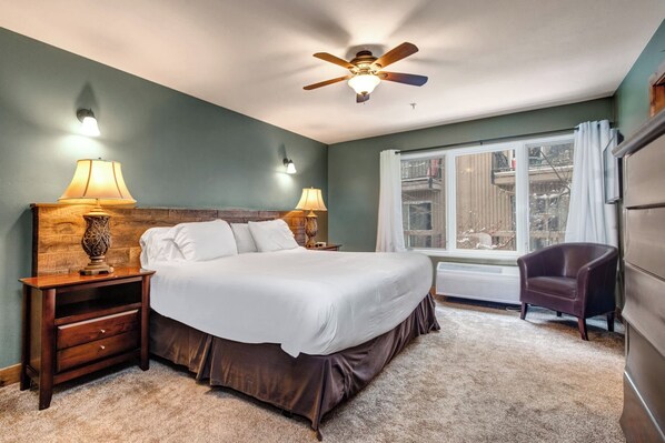 A bedroom with a large bed, two bedside lamps, a ceiling fan, a window with curtains, and a chair.