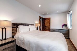 Un dormitorio con una cama, mesitas de noche, cabecera de madera, una ventana con cortinas y una puerta.