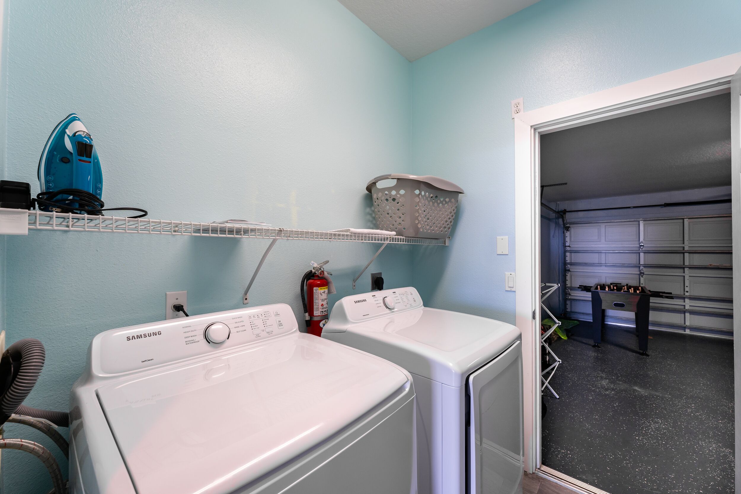 A laundry area with two white washers and dryers, a blue vacuum cleaner, a fire extinguisher, and a basket on a shelf.