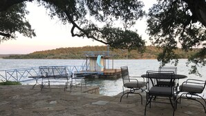 Patio affacciato su un lago, con pavimento in pietra, sedie in metallo e un tavolo. Sullo sfondo, si intravede una struttura per scivoli d'acqua blu.