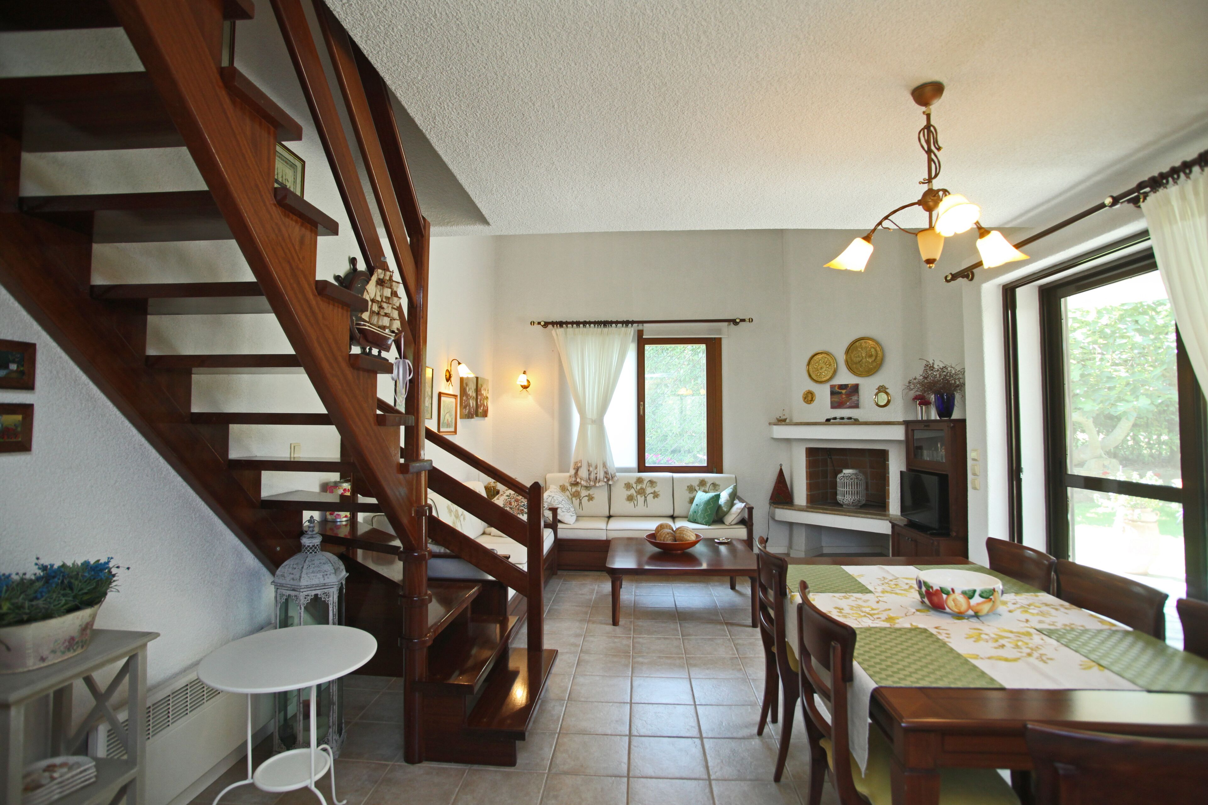 Un salon chaleureux avec un escalier en bois, un coin repas avec une table et des chaises, et une cuisine avec une fenêtre et des rideaux.