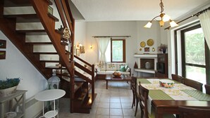 Een gezellige woonkamer met een houten trap, een eethoek met een tafel en stoelen, en een keuken met een raam en gordijnen.