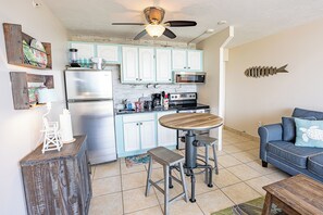 A compact kitchen with a refrigerator, microwave, and stove, a dining area with a wooden table and stools, and a living area with a sofa and decorative pillows.