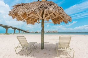 A beach with two metal lounge chairs under a thatched umbrella, a wooden post with a sign, and a bridge in the background.