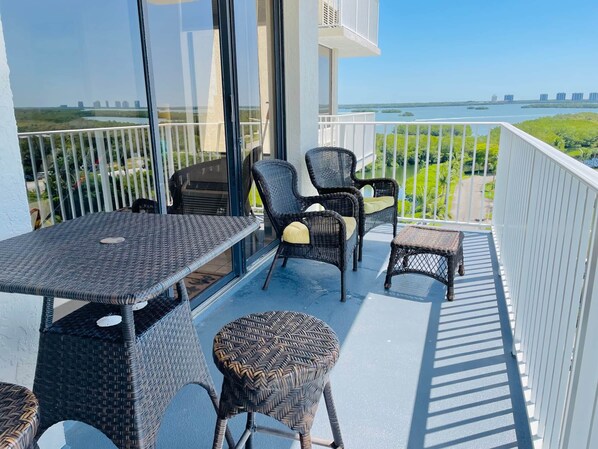 A balcony with wicker furniture, a table, and chairs, overlooking a body of water and greenery.