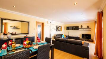 A modern living room with a sofa, a dining area with a table set for two, and a flat-screen TV mounted on the wall.