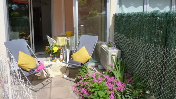 Ein Balkon mit zwei Liegestühlen, einem kleinen Tisch mit gelber Tischdecke und einem Blumentopf mit violetten Blüten.