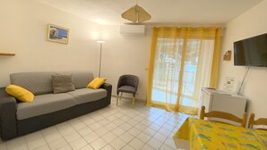 Ein Wohnzimmer mit grauem Sofa, einem kleinen Sessel, einem gelben Tisch mit gemustertem Tischläufer, einem Fernseher an der Wand und einem Fenster mit gelben Vorhängen.