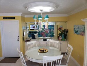 A dining area with a round glass table, white chairs, a chandelier, and a kitchen in the background.