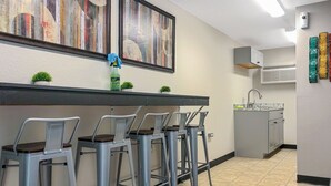Een keuken met een betegelde vloer, een spoelbak en een aanrecht. Er staan vier metalen barkrukken met houten zitplaatsen. Boven het aanrecht hangen twee abstracte schilderijen.