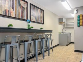 Una cocina con suelo de baldosas, un lavamanos y una encimera. Hay cuatro taburetes de metal con asientos de madera. Dos cuadros abstractos cuelgan de la pared sobre la encimera.