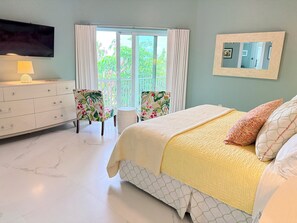 Une chambre à coucher avec un lit, une commode, un téléviseur, une fenêtre avec des rideaux et une chaise.
