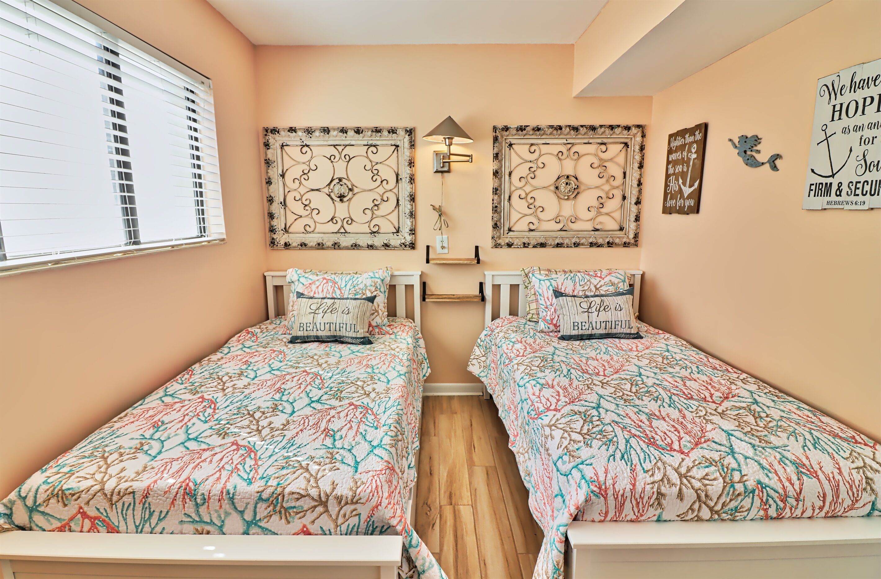 Quarto com duas camas, cada uma com roupa de cama e almofadas estampadas com flores e o texto “Life is Beautiful”. Há quadros decorativos acima das camas e uma luminária fixada na parede.