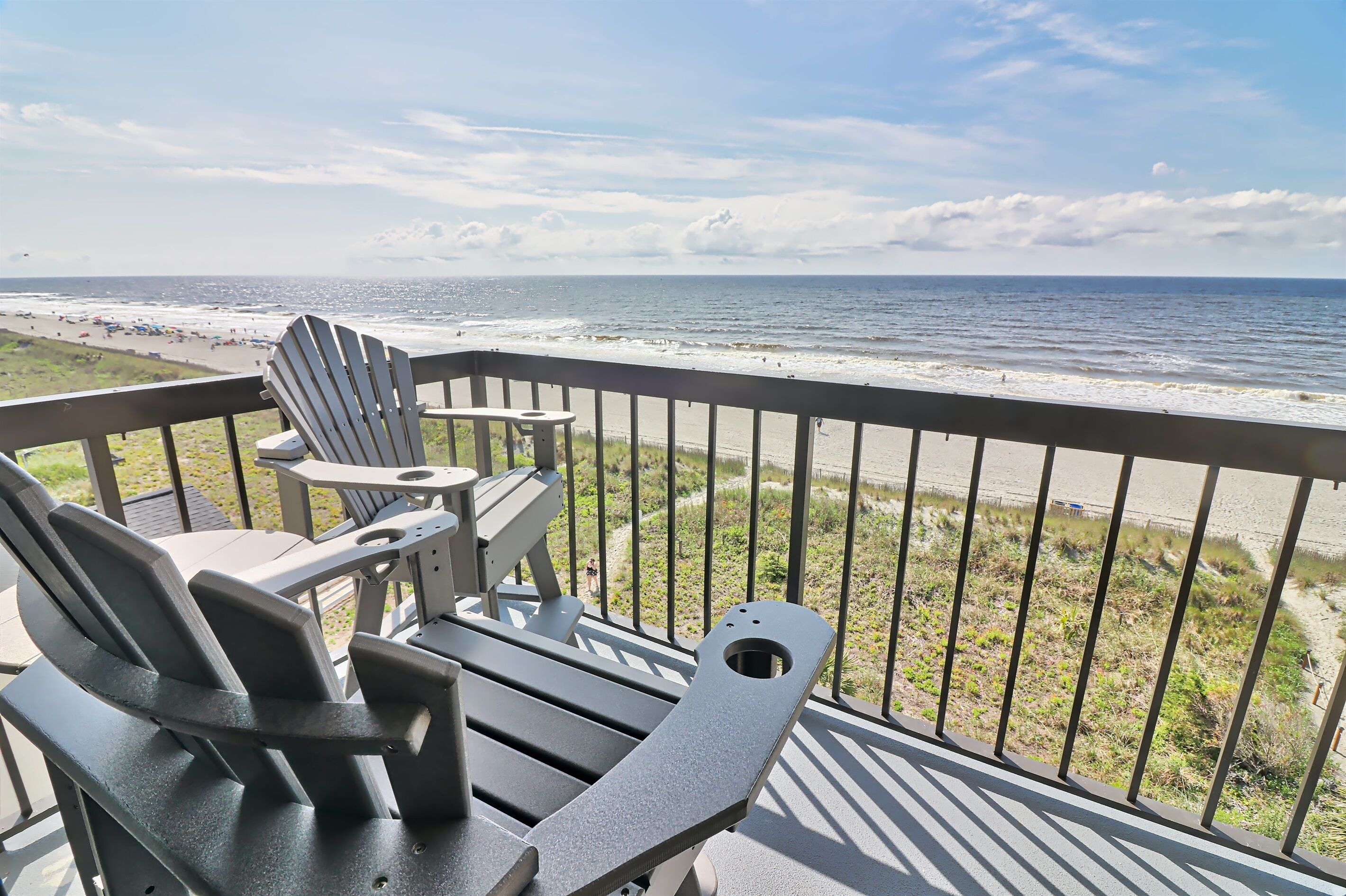 A deck with Adirondack chairs and a small table overlooking a beach.