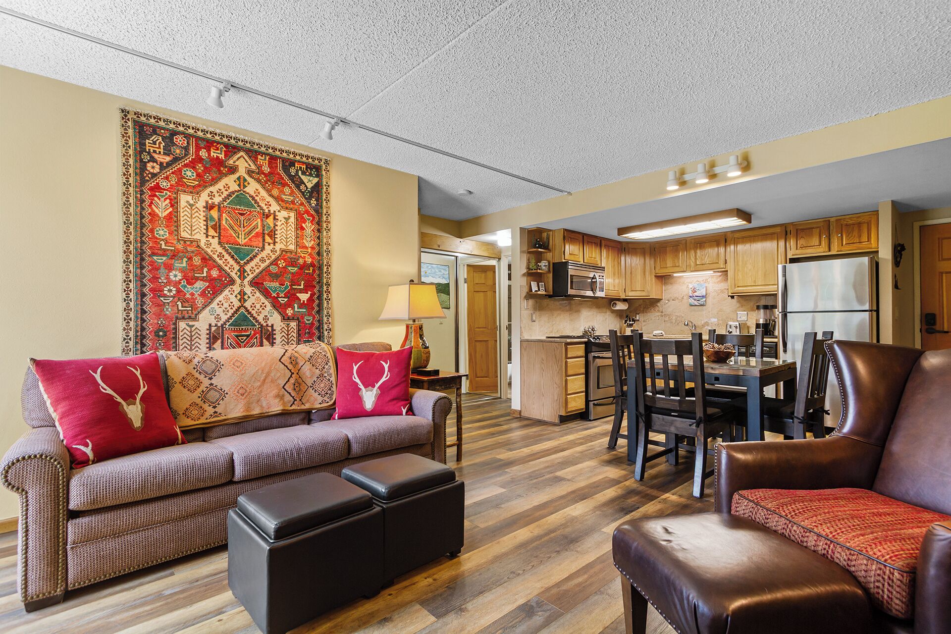 A living room with a sofa, ottoman, and a patterned rug. There is a kitchen area with wooden cabinets and appliances.
