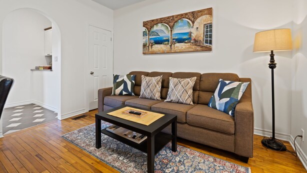 Ein Wohnzimmer mit einem braunen Sofa, gemusterten Kissen, einem Couchtisch, einem Gemälde über dem Sofa und einer Lampe rechts.