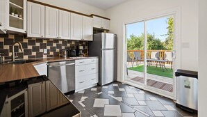 Eine moderne Küche mit weißen Schränken, einem Edelstahl-Kühlschrank und einem Geschirrspüler. Eine Glastür führt auf eine Terrasse mit Holzterrasse und Sitzgelegenheiten.