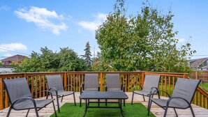 Eine Holzterrasse mit Tisch und vier Stühlen, umgeben von grünem Kunstrasen und einem Hollerzaun.
