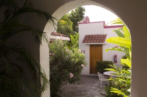 An outdoor courtyard with a white wall, a wooden door, and a tiled roof. There is a small table and chairs, and various plants and flowers.