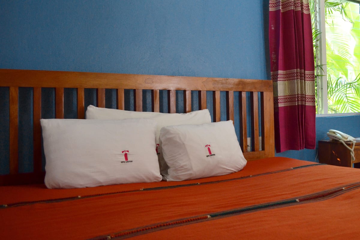 Una cama con cabecera de madera, dos almohadas blancas con un logotipo, una colcha naranja y un teléfono sobre una mesita de noche.