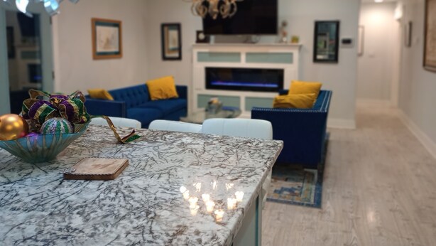 A modern living room with a marble coffee table, blue sofas with yellow cushions, a chandelier, and a flat-screen TV mounted above a fireplace. - Totally Renovated  with Amazing Kitchen, Luxurious Master Suie and Private Pool  (Lake Charles)