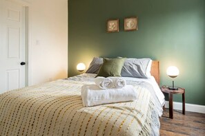 Una cama bien tendida con una toalla enrollada, dos lámparas de noche y un cuadro en la pared.