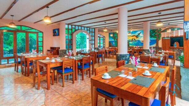 A spacious dining area with wooden tables and chairs, large windows offering a view of greenery, and a high ceiling with exposed beams. - Grand Hotel Leon Marino Galapagos (Puerto Ayora)