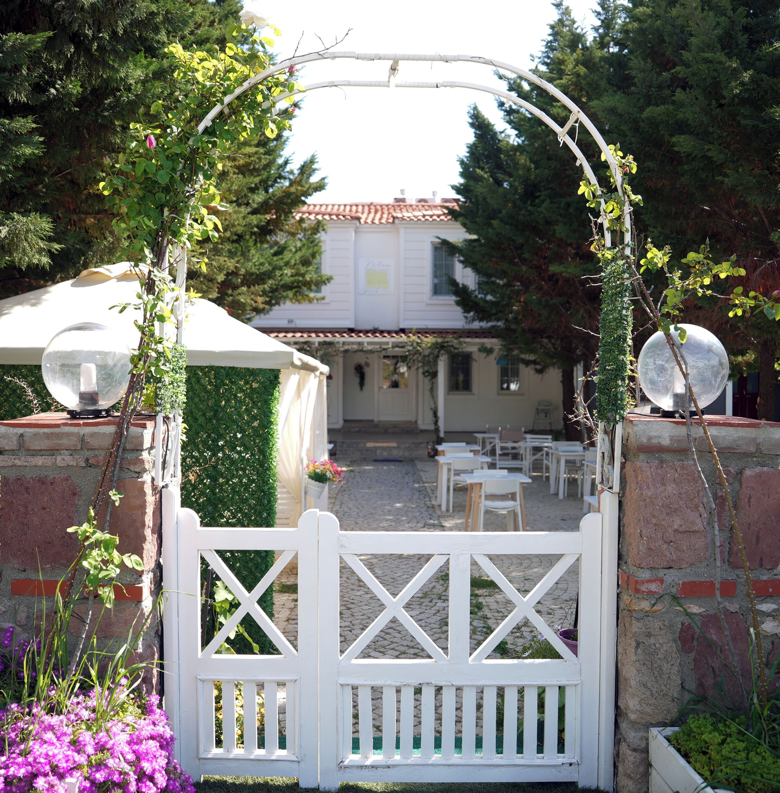 Un portón blanco de madera con un arco decorativo, que da paso a un patio con mobiliario exterior y un edificio al fondo.