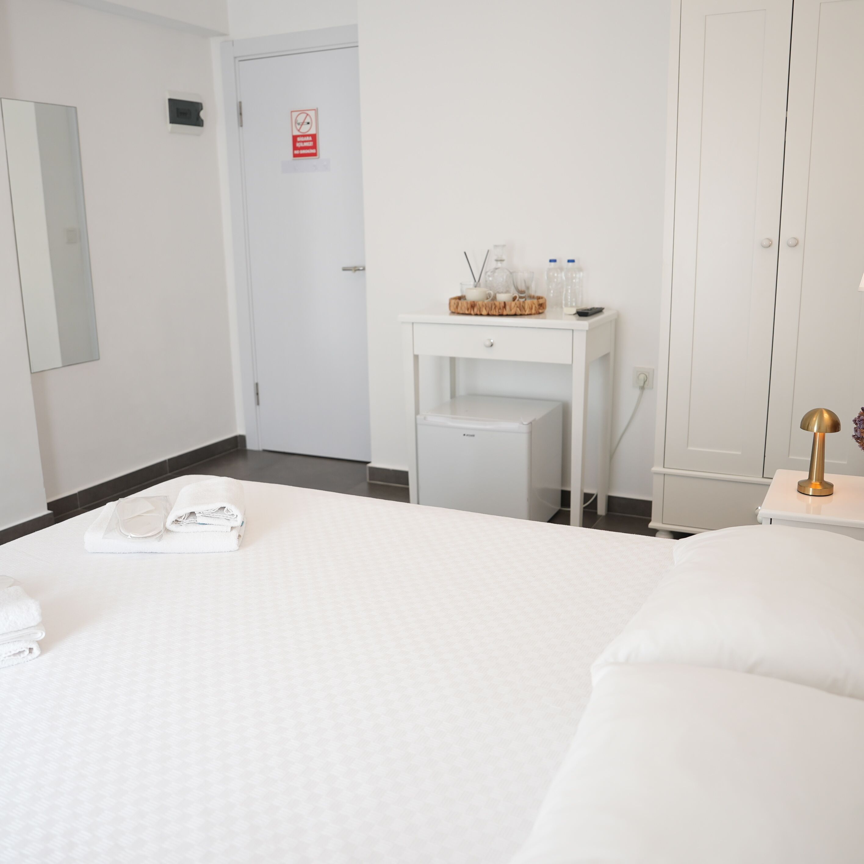 A hotel room with a neatly made bed, a small bedside table with a lamp, a mirror, and a door with a no-smoking sign.