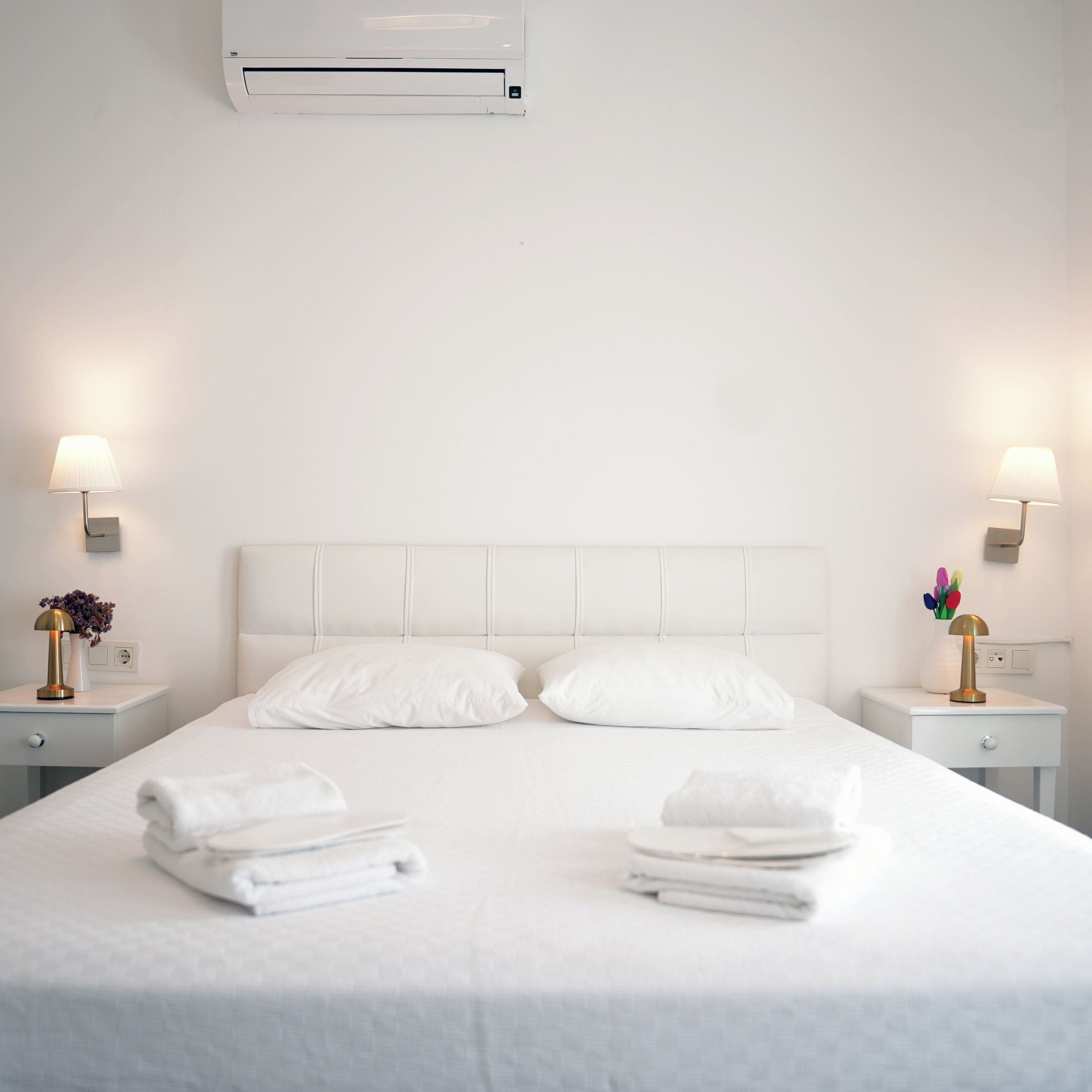 A hotel room with a large bed, two bedside tables with lamps, and an air conditioning unit above the bed.