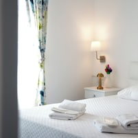 Una cama bien hecha con toallas blancas dobladas, una lámpara de noche y un pequeño florero con flores de colores sobre una mesita blanca.