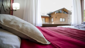 Un letto con una coperta rossa e cuscini bianchi, una finestra con tende e vista su un edificio in legno.