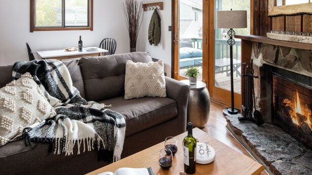 A cozy living room with a fireplace, a sofa, a coffee table with a wine bottle and glasses, and a dining area with a table and chairs. - Outpost: Spruces 10 - Located in the Aspens! (Wilson)