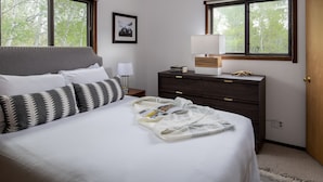 A neatly made bed with a patterned pillow, a bedside table with a lamp, a dresser with a mirror, and a window with a view of trees. - Outpost: Spruces 10 - Located in the Aspens! (Wilson)