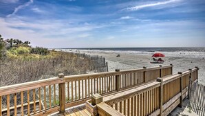Eine Holzterrasse mit Blick auf einen Strand, ein roter Sonnenschirm und Menschen in der Ferne.