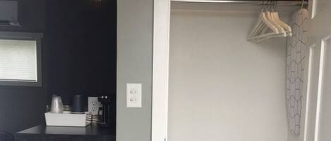 A compact kitchen with a microwave, coffee maker, and a small refrigerator. There is a hanging light fixture above the counter and a door leading to a closet with a hanging rack.