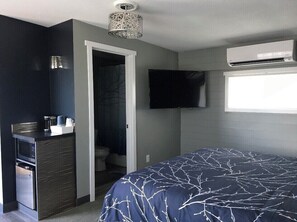 A modern bedroom with a dark blue bedspread, a built-in TV, an air conditioning unit, and a kitchenette area. - #5 King- View of Lake Superior, 2 minute walk to Burlington Bay!  (Two Harbors)