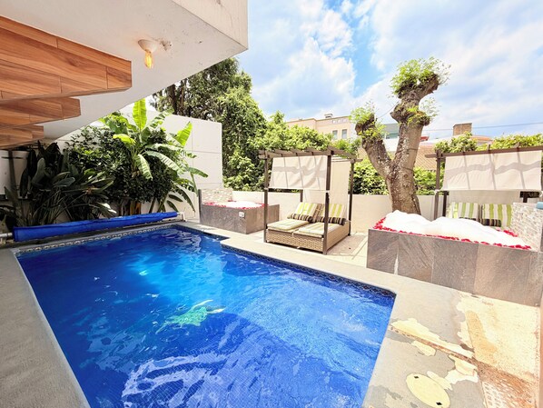 A swimming pool with a sunbed and a pergola, surrounded by greenery and a clear sky.