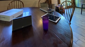 A wooden dining table with a laptop, a tablet, and a purple cup. There are chairs around the table, and a large window with blinds is visible. - One of a kind cozy Renovated VT Barn- where Family, Friends, Couples gather  (Shaftsbury)