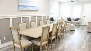 A dining area with a wooden table and chairs, a gray wall with a painting, a ceiling fan, and a gray couch in the background.
