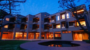 Un immeuble à plusieurs étages, doté de balcons et de grandes fenêtres, entouré d’un espace paysager agrémenté d’un élément décoratif aquatique circulaire.