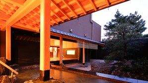 Un onsen japonais traditionnel, avec une structure en bois, une source d’eau chaude entourée de pierres et une vue sur un pin.