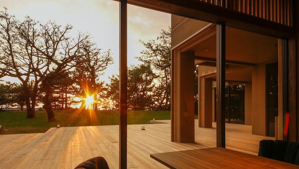Un espace extérieur moderne, aménagé pour se détendre, avec une terrasse en bois, une table et des chaises, offrant une vue sur un paysage verdoyant, parsemé d’arbres, et sur un coucher de soleil.
