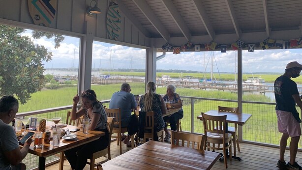 A covered outdoor seating area with tables and chairs, overlooking a body of water and farmland.