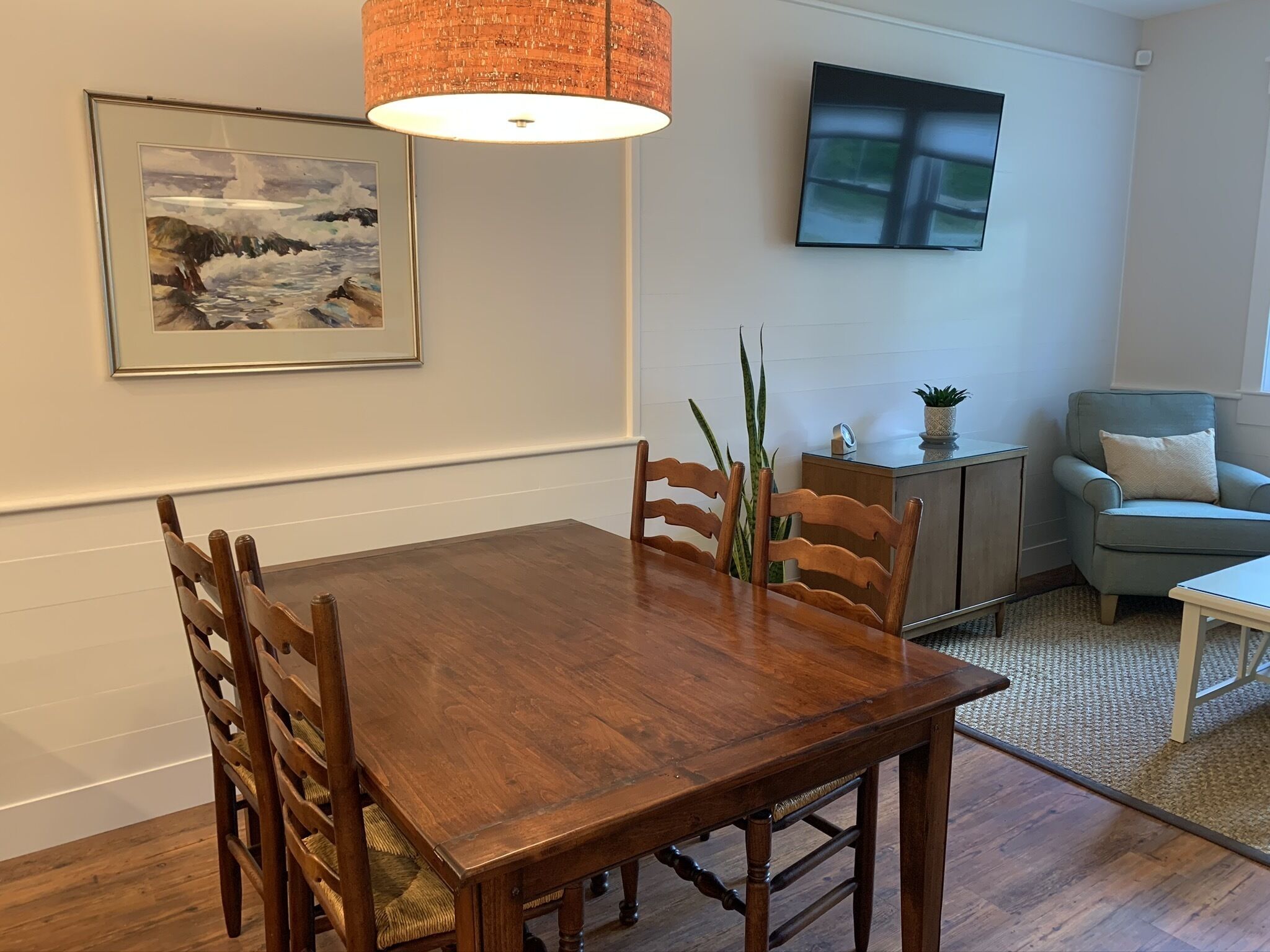 A dining area with a wooden table and chairs, a hanging lamp, a framed painting, a television, and a sofa.