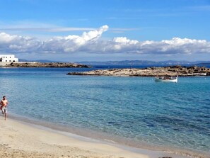 A beach with clear blue water, a rocky outcrop, and a white building in the background.