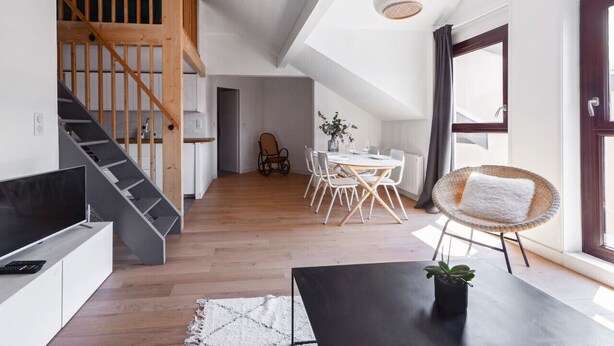 Un appartement moderne de style « loft », doté d’un escalier en bois, d’un coin repas avec une table et des chaises blanches, et d’une grande fenêtre agrémentée de rideaux.